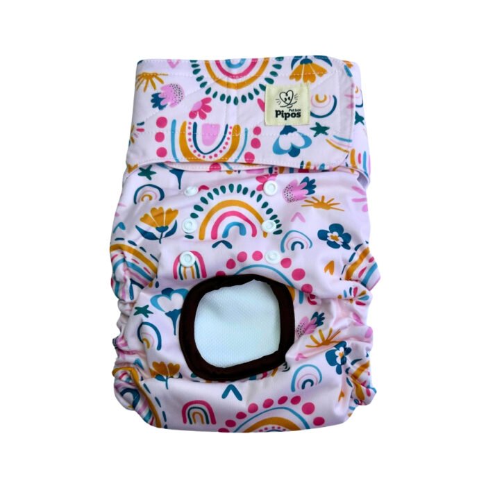 Vista frontal del pañal reutilizable para perrita, modelo Arco de Sol, con estampado de arcoíris en tonos mostaza, rosa y azul, junto a flores y formas decorativas sobre fondo rosa