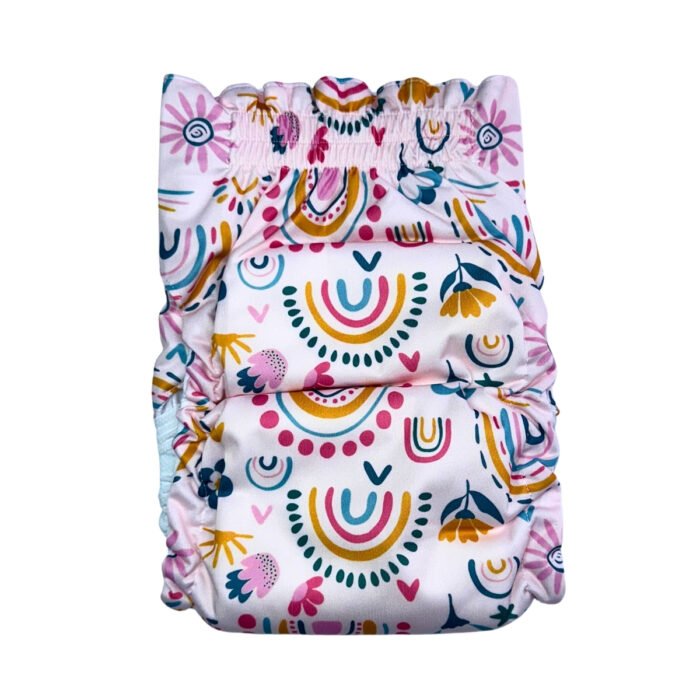Vista trasera del pañal reutilizable para perrita, modelo Arco de Sol, con estampado de arcoíris en tonos mostaza, rosa y azul, junto a flores y formas decorativas sobre fondo rosa